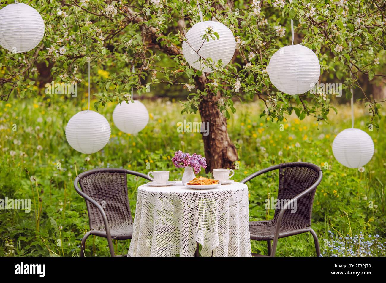 Décoration mignonne avec meubles de jardin, deux tasses à thé et gâteau sur une table. Lanternes en papier chinois suspendues à un pommier en fleur pour la décoration. Banque D'Images
