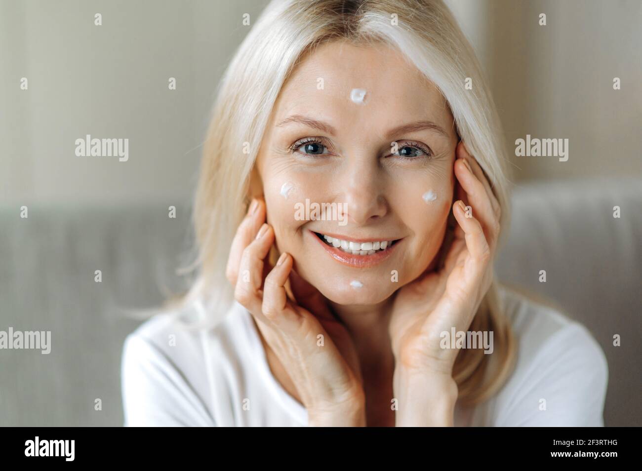 Belle femme blonde caucasienne senior appliquant de la crème anti-âge sur  son visage tout en étant assise sur le canapé à la maison dans le salon,  regardant la caméra et souriant. Concept