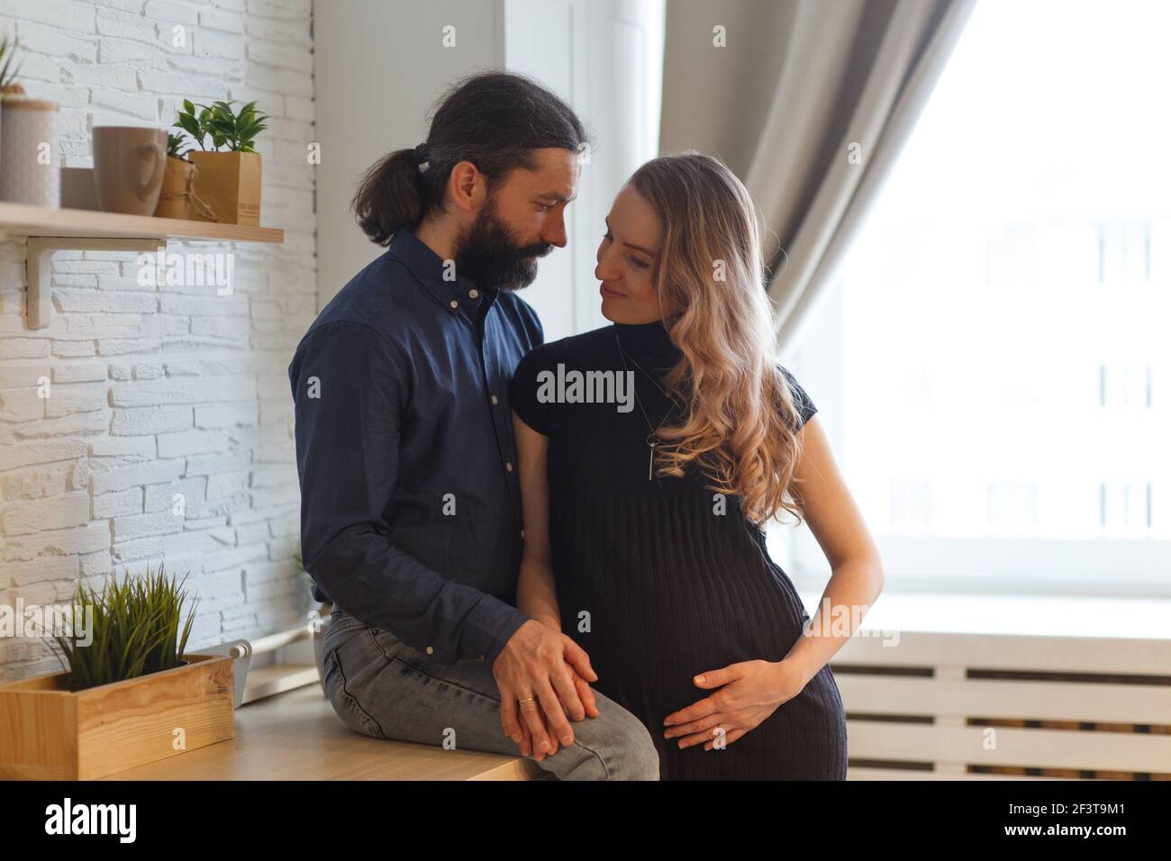 Homme embrassant partenaire enceinte dans la cuisine. Concept de grossesse familiale adulte. Les futurs parents dans la tenue à la maison embrassent debout dans la cuisine, regardant e Banque D'Images