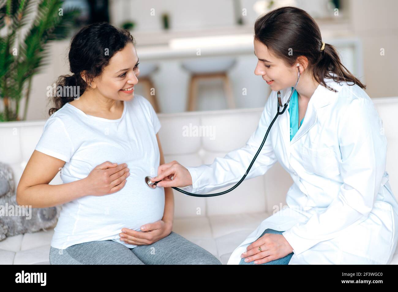 Sante De La Femme Enceinte Et Du Bebe Femme Sympathique Medecin Examine La Femme Souriante De Race Mixte Enceinte A L Ecoute Des Battements De Cœur De Bebe Assis Sur Un Canape A