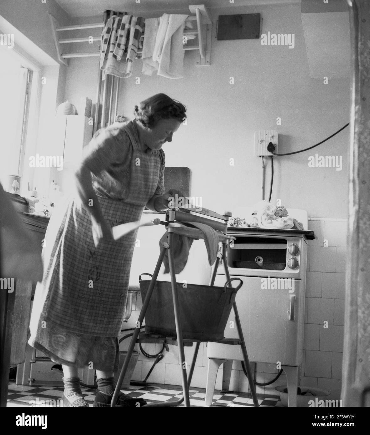 1950s, historique, une femme mûre dans une pinafore dans une cuisine utilisant un simple mangle pour extraire l'eau des vêtements lavés, Angleterre, Royaume-Uni, avec un seau en métal en dessous pour capturer l'eau en excès. Une mangle ou un essoreur, comme on l'appelle également, est une aide à la lessive mécanique, qui a deux rouleaux dans un cadre et est travaillé par une manivelle pour essorer l'eau de la lessive humide. Banque D'Images