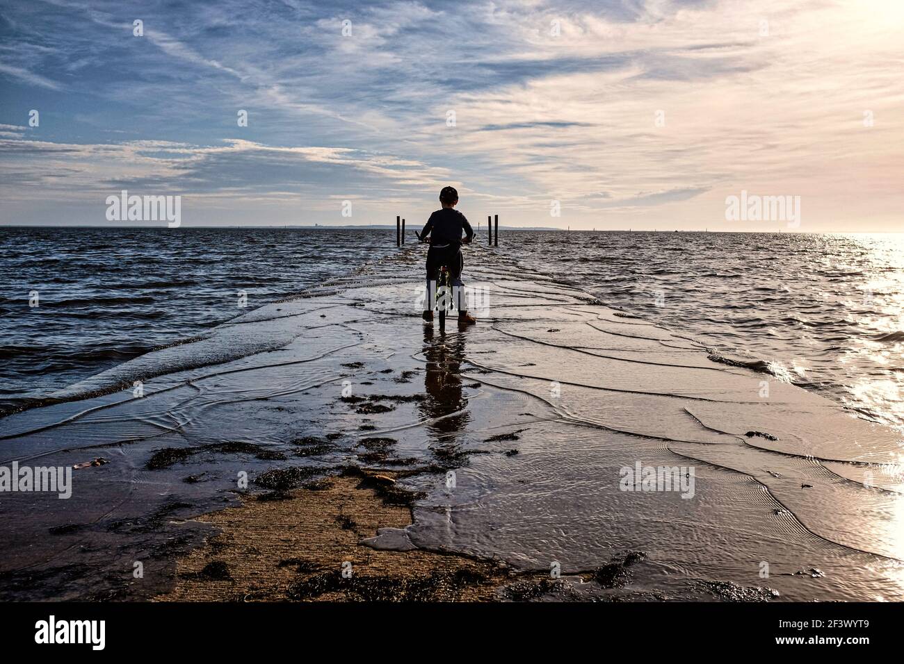 Ares (sud-ouest de la France) : ambiance nocturne au crépuscule et au coucher du soleil sur la baie d'Arcachon en automne. Enfant sur son vélo, ses pieds dans l'eau, face à t Banque D'Images