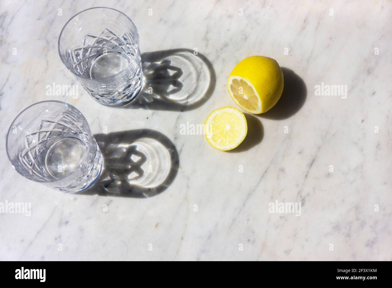 Deux verres d'eau et un citron dans la lumière du soleil avec de longues ombres sur un fond texturé de marbre gris et blanc vue de dessus vers le bas et mise au point intentionnelle. Banque D'Images