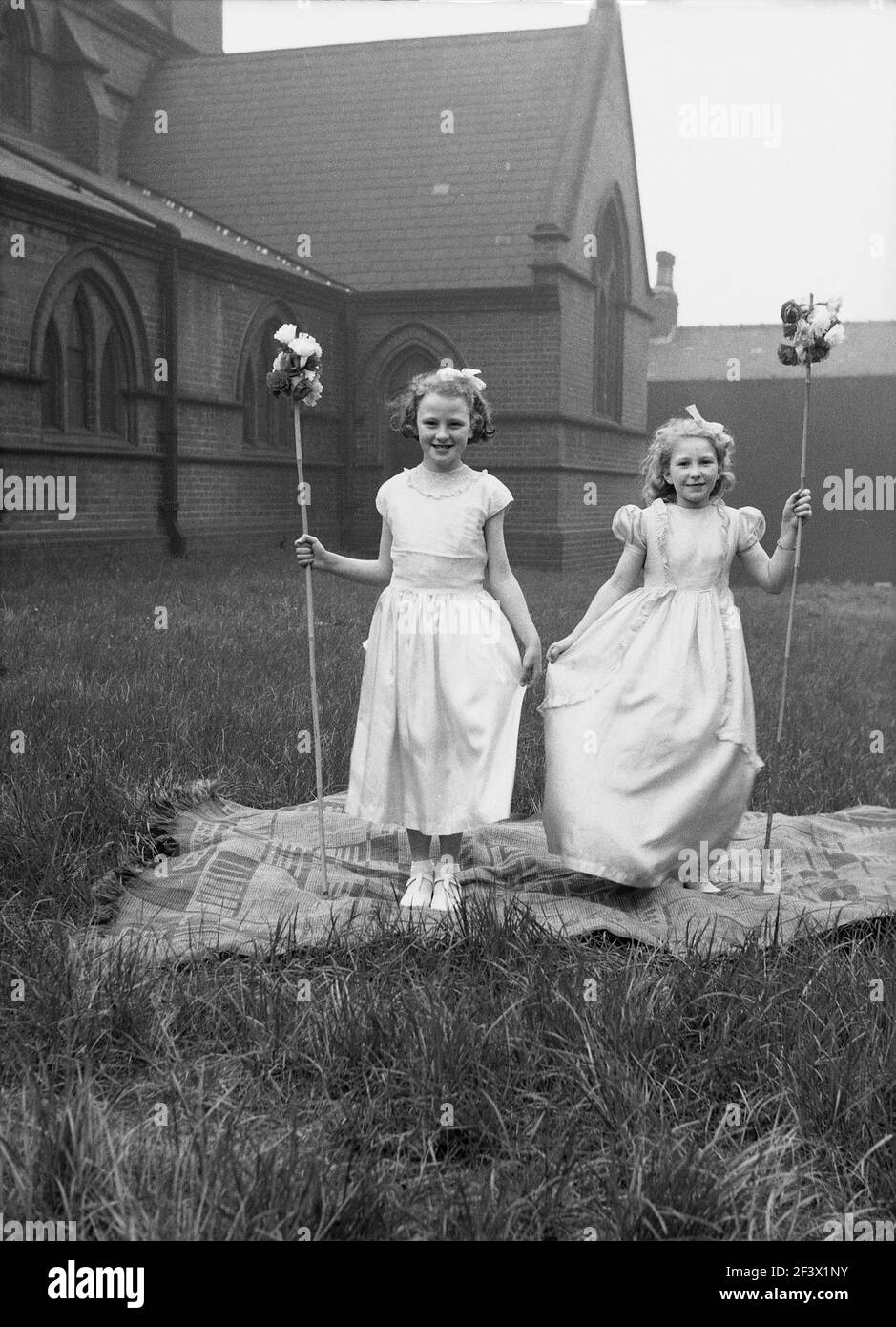 1958, historique, printemps et deux jeunes filles dans leurs costumes et tenant des bâtons de fleurs debout sur un tapis dans le terrain d'une église pour leur photo, avant de prendre part à la parade et au festival du jour de mai, Leeds, Angleterre, Royaume-Uni. Banque D'Images