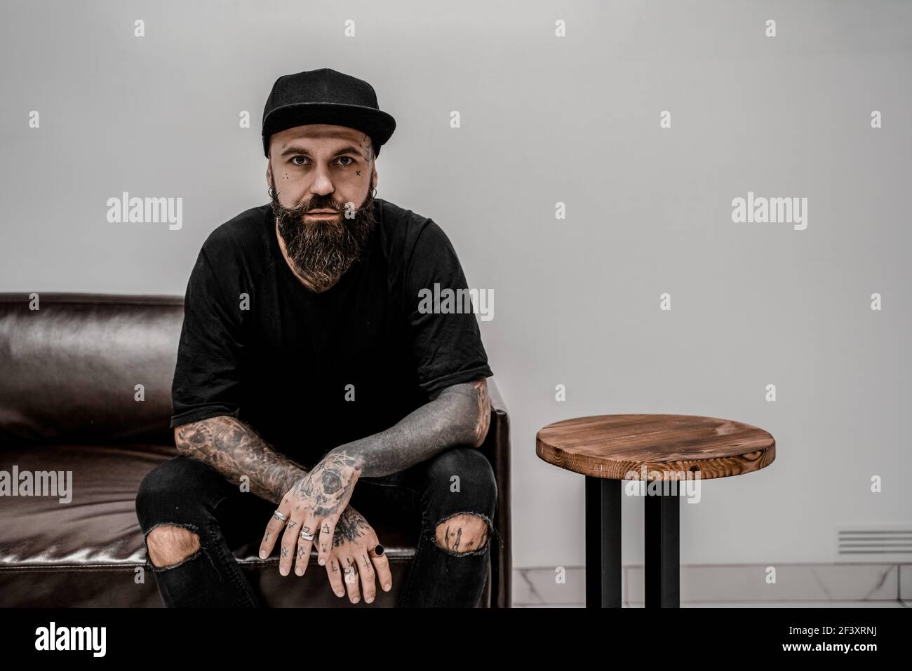 Un jeune artiste de tatouage masculin avec une barbe a l'air sérieux assis sur un canapé dans un lieu d'atelier. Banque D'Images