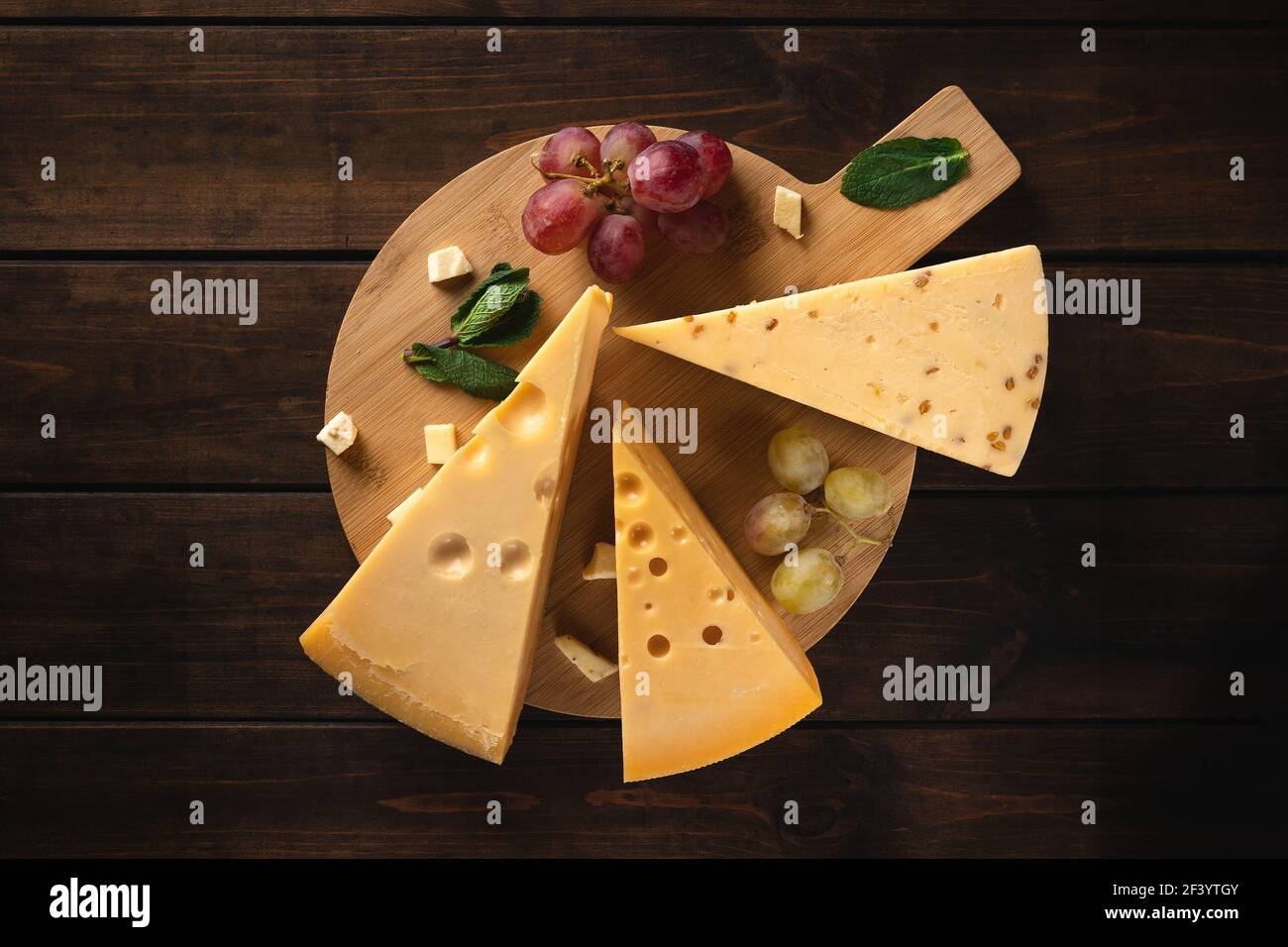 trois morceaux de fromage suisse jaune avec trous et une branche de raisin rouge sur une planche à découper sur fond de bois, vue de dessus Banque D'Images