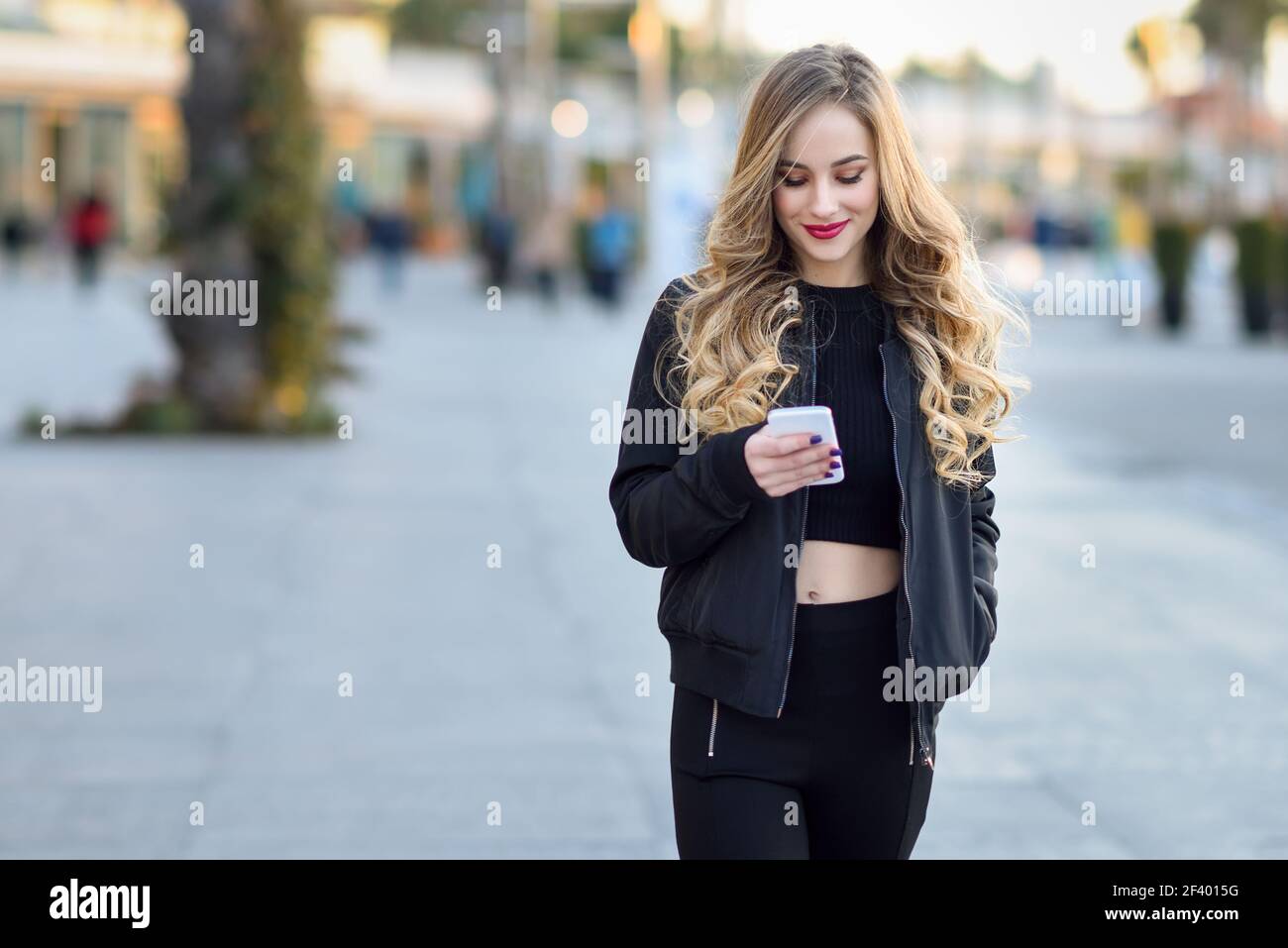 Femme blonde envoyant des SMS avec son smartphone en milieu urbain. Une femme blonde envoie des SMS avec son smartphone en milieu urbain. Belle jeune fille souriante portant une veste noire marchant dans la rue. Jolie femme russe avec une longue coiffure ondulée. Banque D'Images