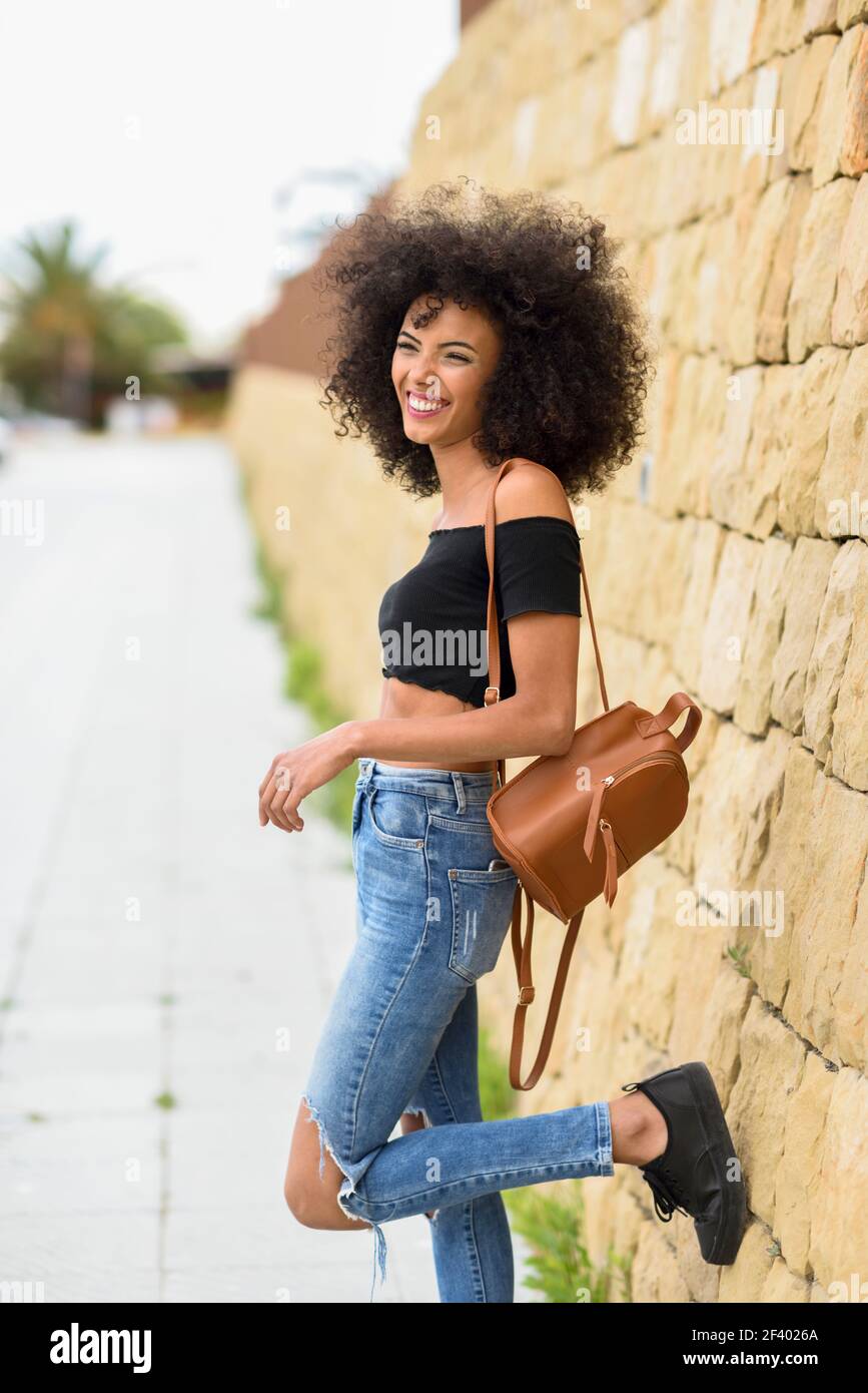 Femme mixte souriante avec cheveux afro debout dans la rue. Bonne femme mixte avec des cheveux afro debout à l'extérieur. Femme portant des vêtements décontractés en milieu urbain. Concept de style de vie Banque D'Images