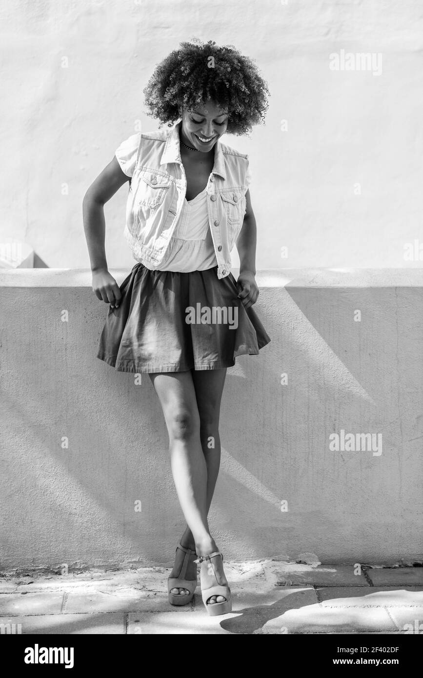 Jeune femme noire, coiffure afro, debout en milieu urbain. Jeune femme noire, coiffure afro, debout dans la rue. Fille portant des vêtements décontractés souriant en milieu urbain. Femme avec jupe, gilet en denim et talons hauts. Banque D'Images