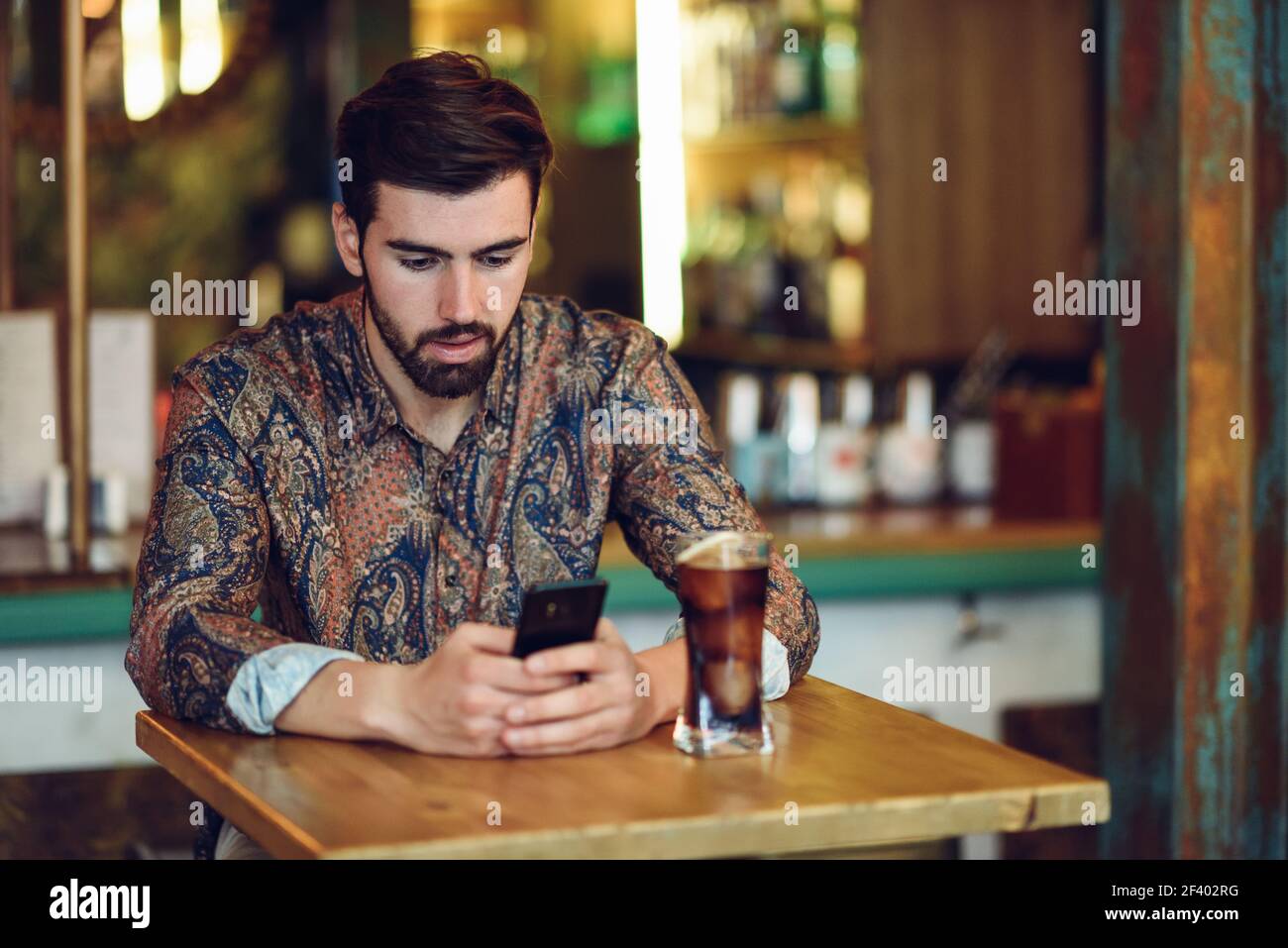 Jeune homme barbu portant des vêtements décontractés en regardant son smartphone dans un pub moderne. Guy avec barbe et coiffure moderne de boire un cola. Banque D'Images