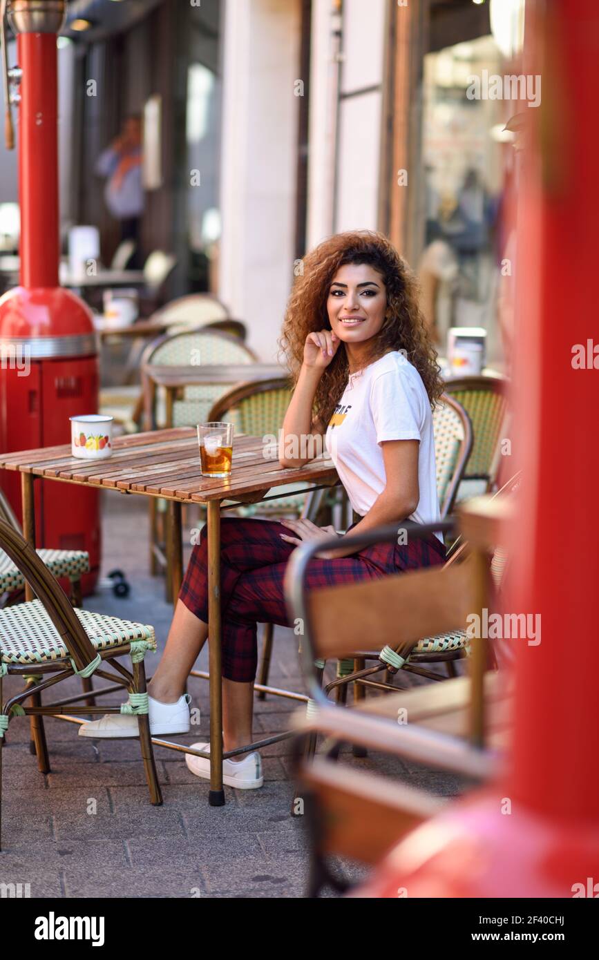 Jeune femme arabe et souriant assis dans un bar urbain dans la rue. Fille arabe dans les tenues de boire un soda à l'extérieur. Banque D'Images