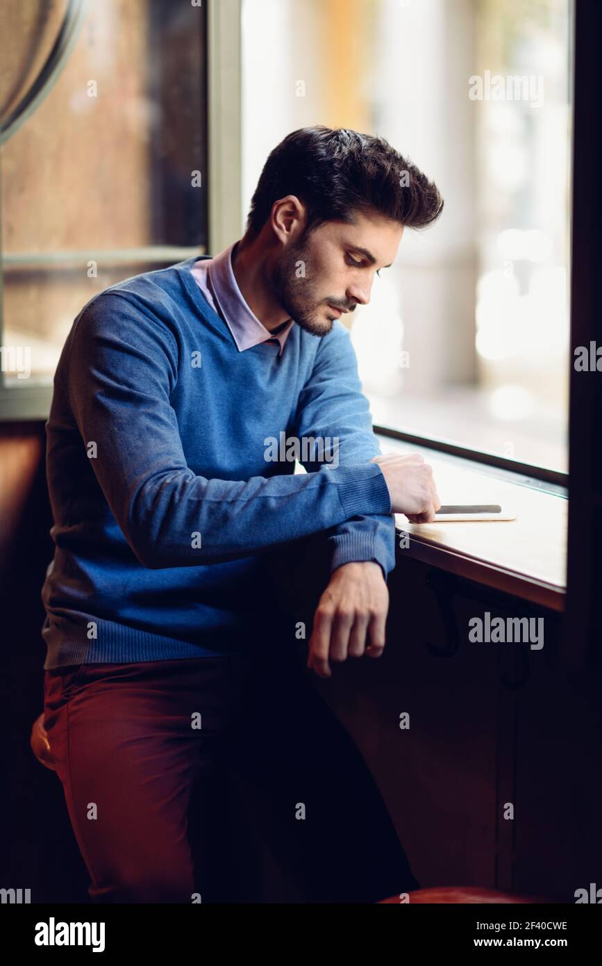 Jeune homme au pull bleu en regardant son smartphone dans un pub moderne. Avec barbu coiffure moderne près d'une fenêtre. Banque D'Images