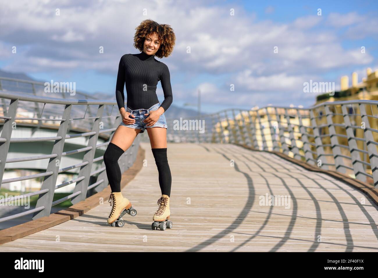 Femme noire, coiffure afro, équitation sur patins à l'extérieur sur le pont urbain à bras ouverts. Souriante jeune femme à roues alignées sur un jour ensoleillé. De beaux nuages dans le ciel. Banque D'Images