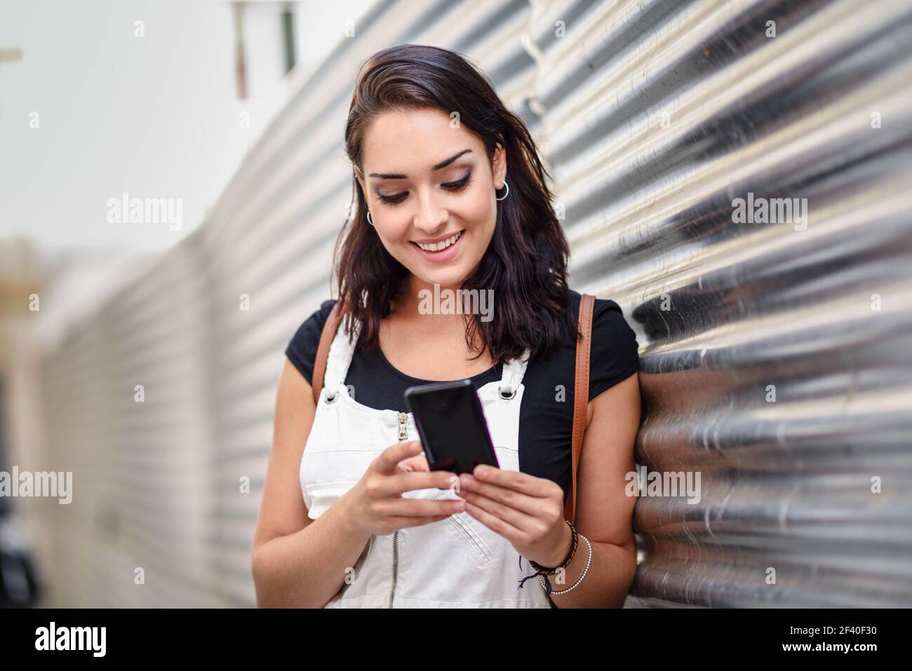 Souriante jeune femme textos avec son téléphone intelligent à l'extérieur. Girl wearing denim dress en contexte urbain. Technologie concept. Banque D'Images