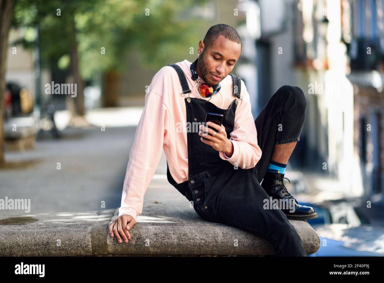 Jeune homme noir avec un casque assis dans la rue en milieu urbain en regardant son téléphone intelligent. Concept de vie. Banque D'Images