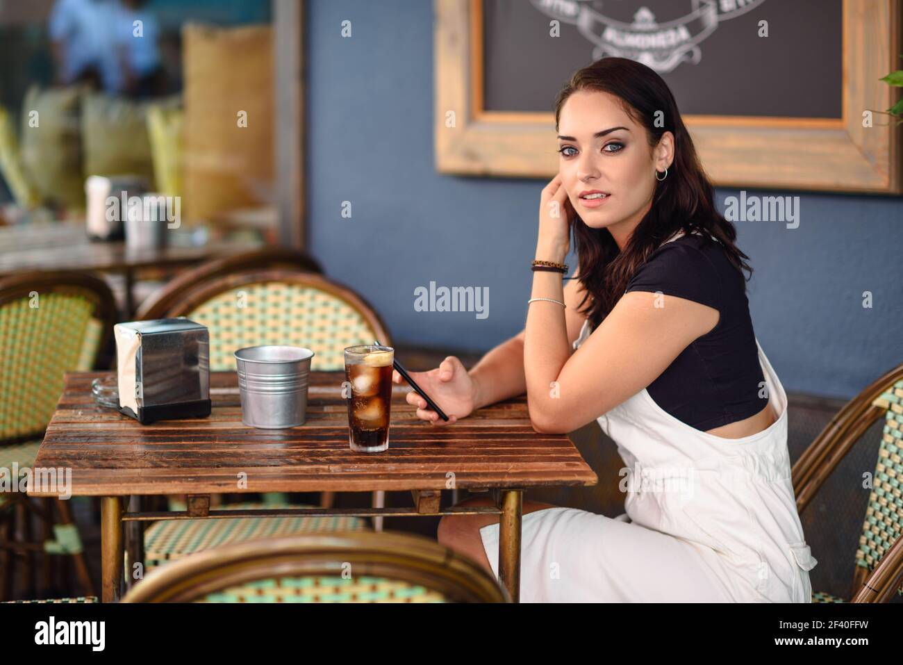 Belle fille aux yeux bleus assis sur un café urbain à l'aide d'un smartphone. Jeune femme à la coiffure ondulée brune portant une robe en denim blanc. Concept de style de vie. Banque D'Images