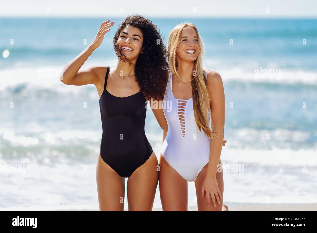 Deux jeunes femmes en maillots de bain sur une plage tropicale. Drôles  caucasiennes et arabes portant des maillots de bain noirs et blancs à  genoux sur la plage. Deux jeunes femmes avec
