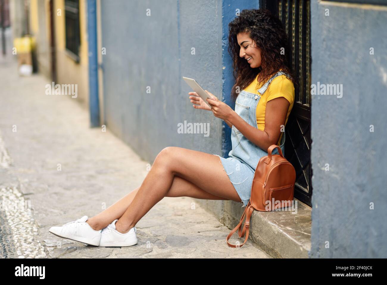 Jeune femme arabe regardant sa tablette numérique à l'extérieur. Fille africaine portant des vêtements décontractés en arrière-plan urbain. Coiffure curly.. Jeune femme arabe regardant sa tablette numérique à l'extérieur. Banque D'Images