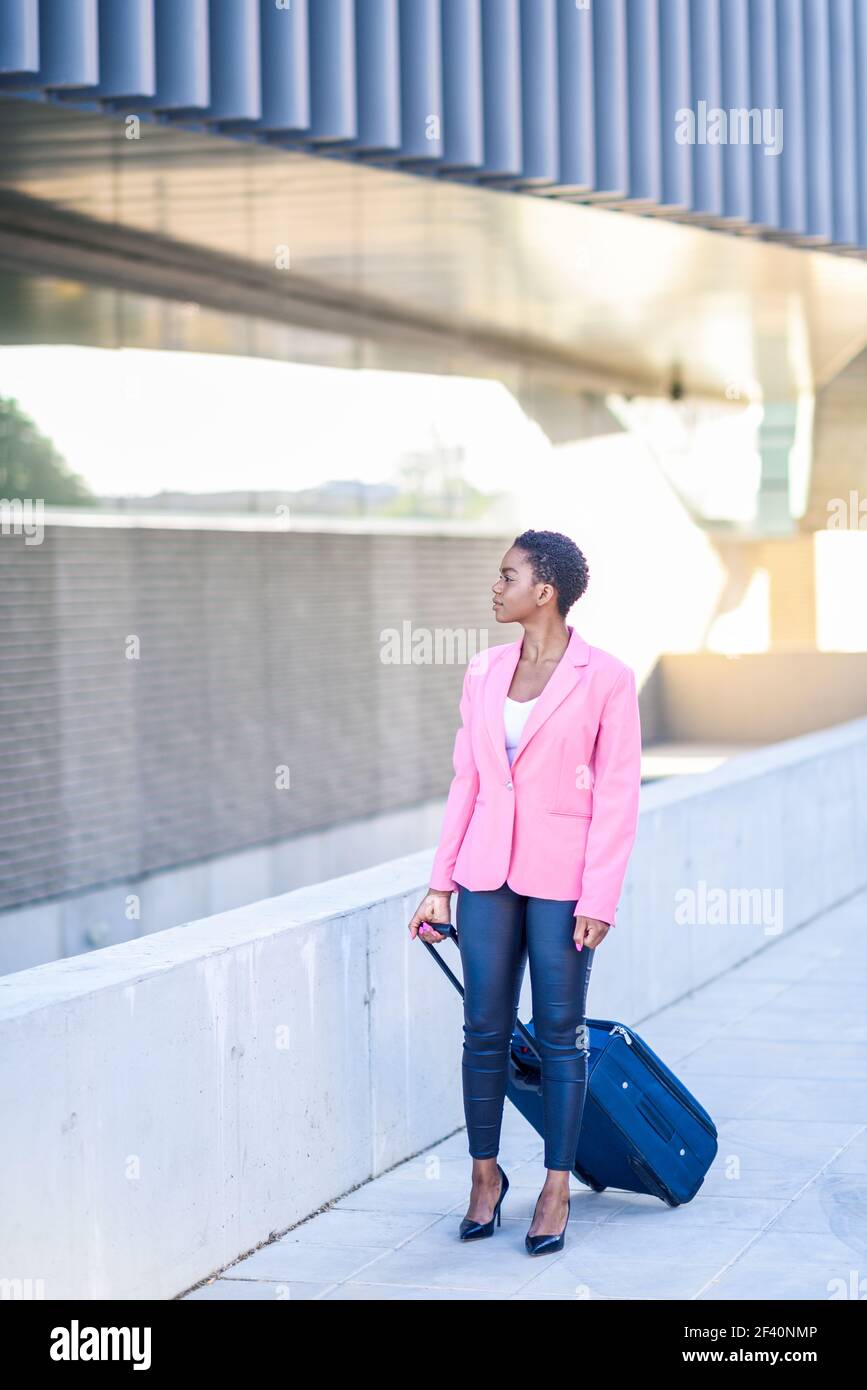 Une femme d'affaires afro-américaine se trouve dans une valise à roulettes près du bâtiment des bureaux d'affaires. Femme noire marchant avec un sac de voyage portant une veste rose. Femme noire marchant avec un sac de voyage portant une veste rose. Banque D'Images