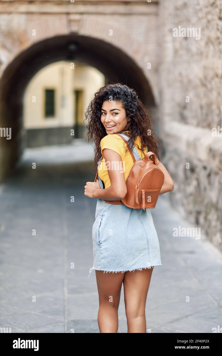 Vue arrière de la jeune femme arabe avec sac à dos à l'extérieur. Fille de voyageur dans des vêtements décontractés dans la rue. Bonne femme portant un t-shirt jaune et une robe en Jean sur fond urbain. Jeune femme nord-africaine avec une coiffure noire en plein air. Banque D'Images