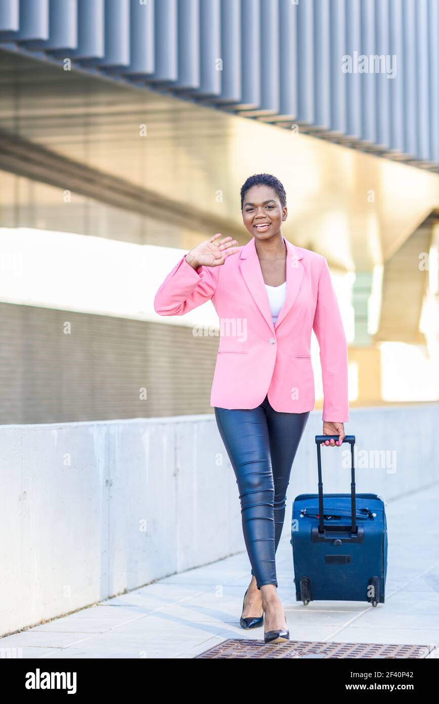 Une femme d'affaires afro-américaine se trouve dans une valise à roulettes près du bâtiment des bureaux d'affaires. Femme noire marchant avec un sac de voyage portant une veste rose. Femme noire marchant avec un sac de voyage portant une veste rose. Banque D'Images
