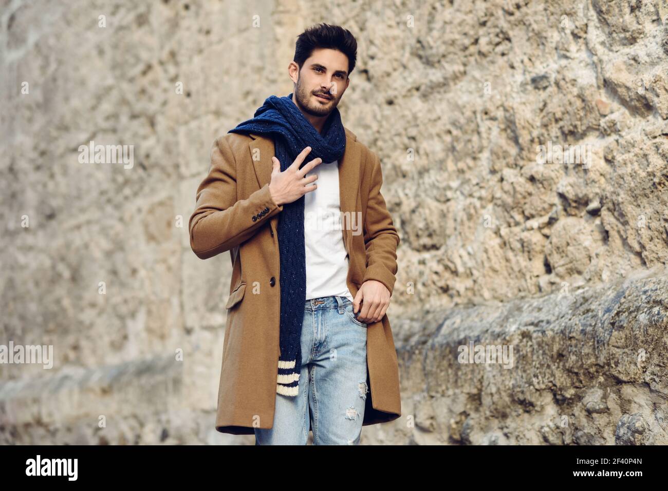 Jeune homme portant des vêtements d'hiver dans la rue. Jeune homme barbu avec une coiffure moderne avec un manteau, une écharpe, un Jean bleu et un t-shirt. Jeune homme portant des vêtements d'hiver dans la rue. Banque D'Images