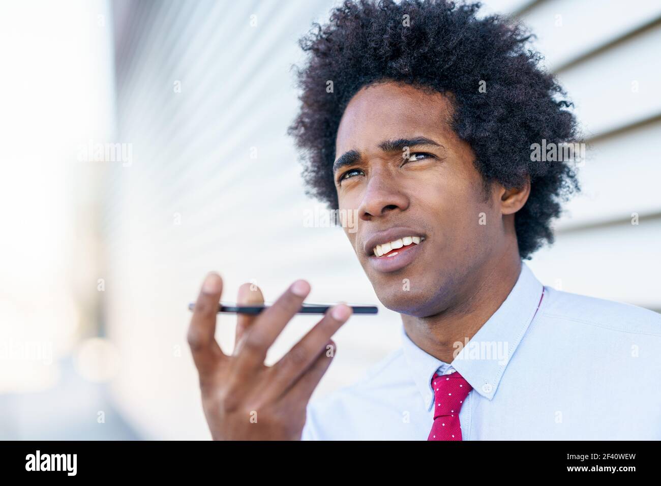 Homme d'affaires noir envoyant une note vocale avec son smartphone près d'un immeuble de bureaux. Homme avec cheveux afro.. Homme d'affaires noir utilisant un smartphone près d'un immeuble de bureaux Banque D'Images