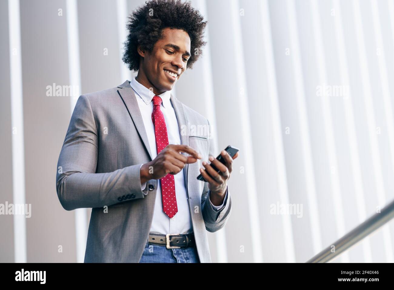 Homme d'affaires noir souriant utilisant son smartphone près d'un immeuble de bureau. Homme avec cheveux afro.. Homme d'affaires noir utilisant un smartphone près d'un immeuble de bureaux Banque D'Images
