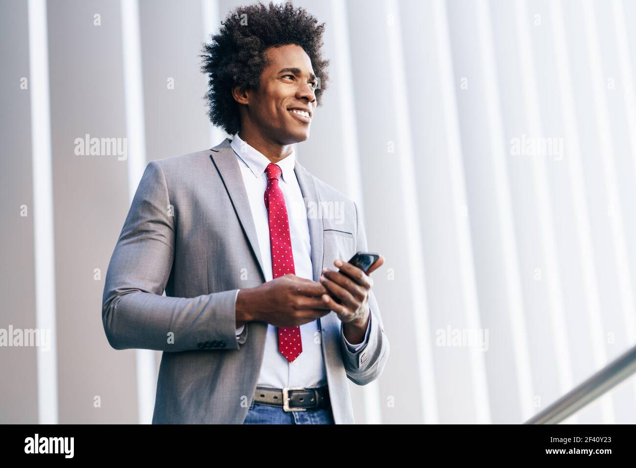 Homme d'affaires noir souriant utilisant son smartphone près d'un immeuble de bureau. Homme avec cheveux afro.. Homme d'affaires noir utilisant un smartphone près d'un immeuble de bureaux Banque D'Images