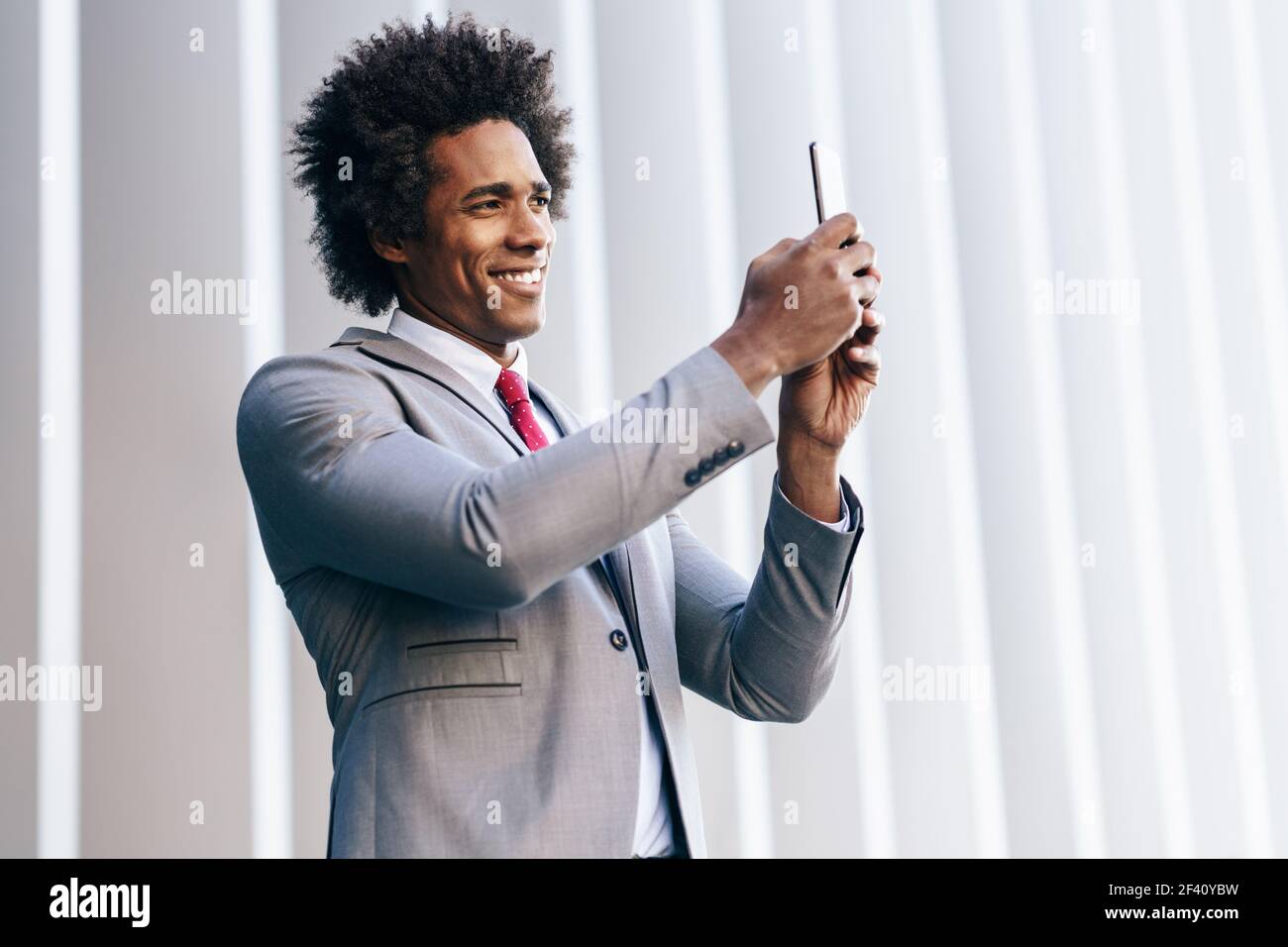 Homme d'affaires noir prenant des photos avec son smartphone près d'un immeuble de bureaux. Homme avec cheveux afro.. Homme d'affaires noir utilisant un smartphone près d'un immeuble de bureaux Banque D'Images