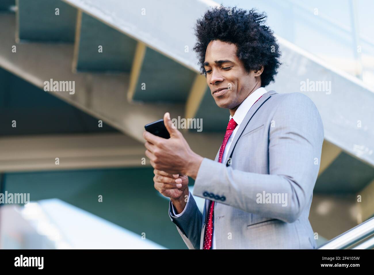 Homme d'affaires noir utilisant son smartphone près d'un immeuble de bureaux. Homme avec cheveux afro.. Homme d'affaires noir utilisant un smartphone près d'un immeuble de bureaux Banque D'Images