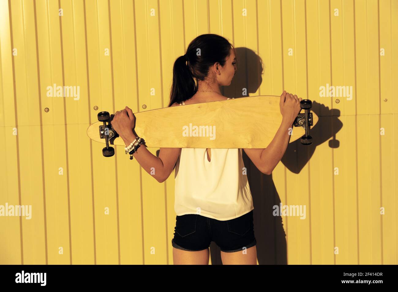 Jeune fille posant avec son skateboard derrière son dos en face de la clôture jaune avec l'espace d'imitation, vue arrière tourné. Jeune fille posant avec son skateboard derrière son dos en face de la clôture jaune avec l'espace d'imitation, vue arrière Banque D'Images