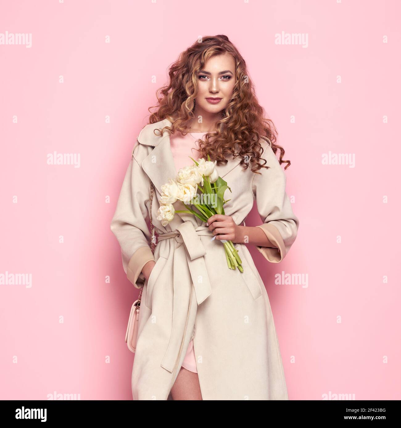 Jeune femme élégante en manteau blanc tendance. Cheveux blonds, robe corail, isolé sur fond rose, prise de vue en studio. Livre de recherche de printemps à la mode. Femme modèle avec sac à main Banque D'Images