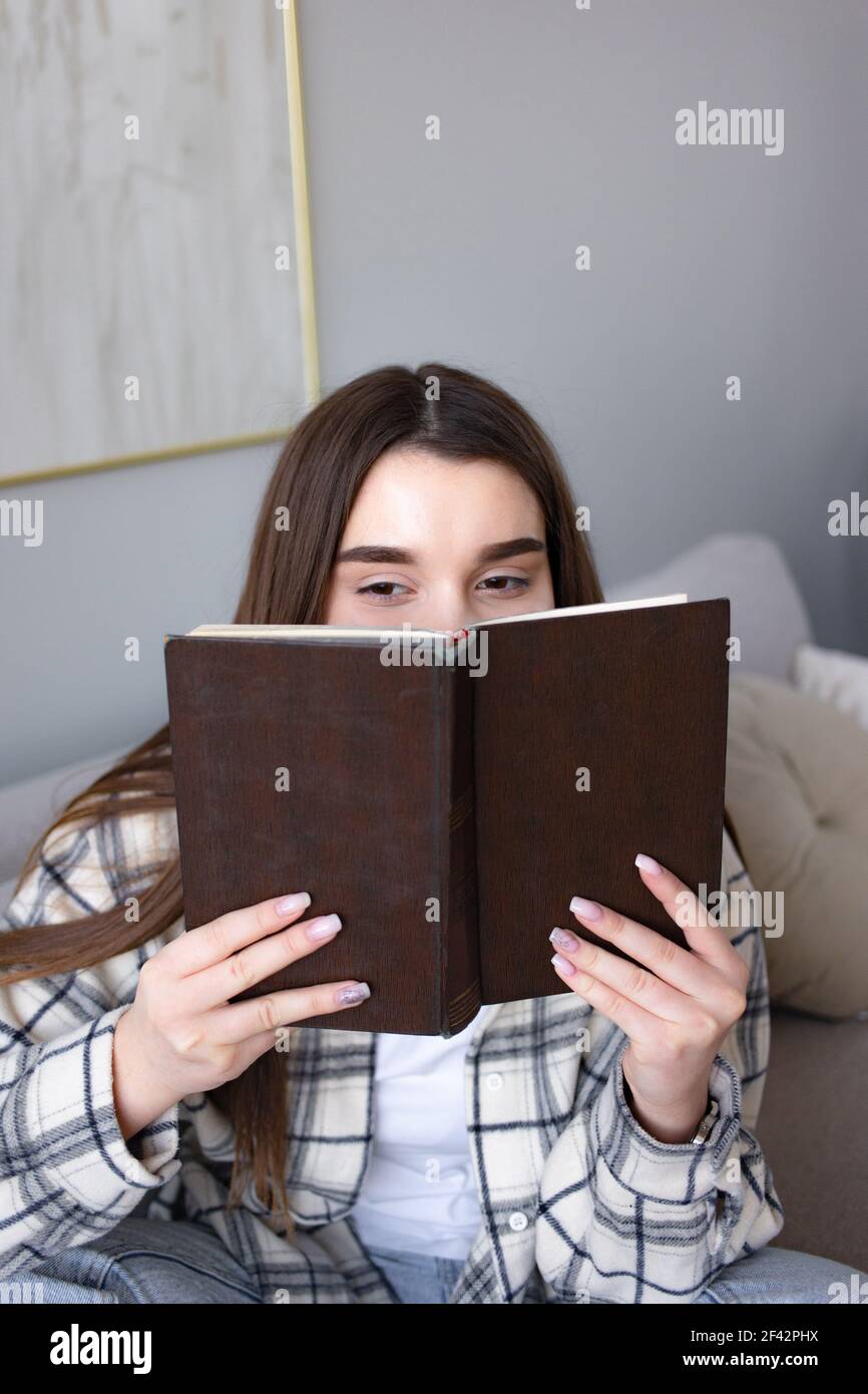 Une femme joyeuse se cache face à un vieux livre, a une expression joyeuse, étant dans l'esprit de la lecture de son histoire préférée, aime la fiction, passe des loisirs avec Banque D'Images