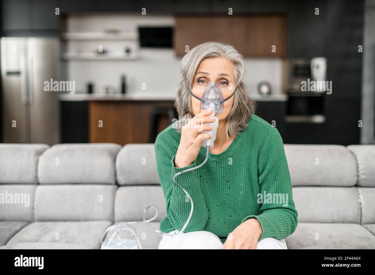 Malade sénior sénior assis sur le canapé dans un salon, portant un masque à oxygène pour traiter la grippe, la toux, le coronavirus, l'inhalation par un inhalateur, se sentant mal et fatigué Banque D'Images