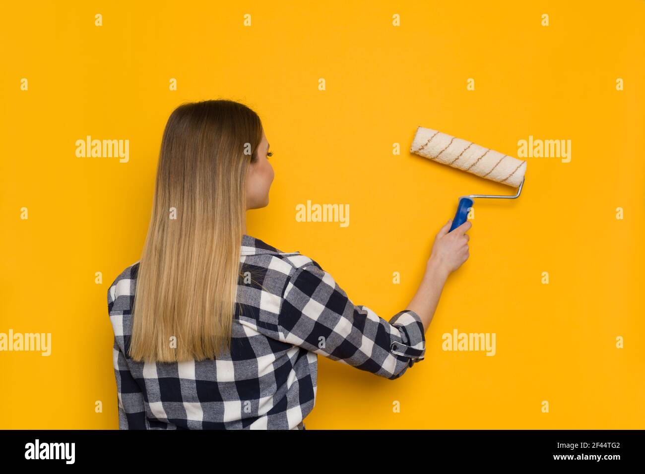 La jeune femme en chemise de bûcherons peint un mur jaune avec un rouleau de peinture, vue arrière. Taille haute. Banque D'Images