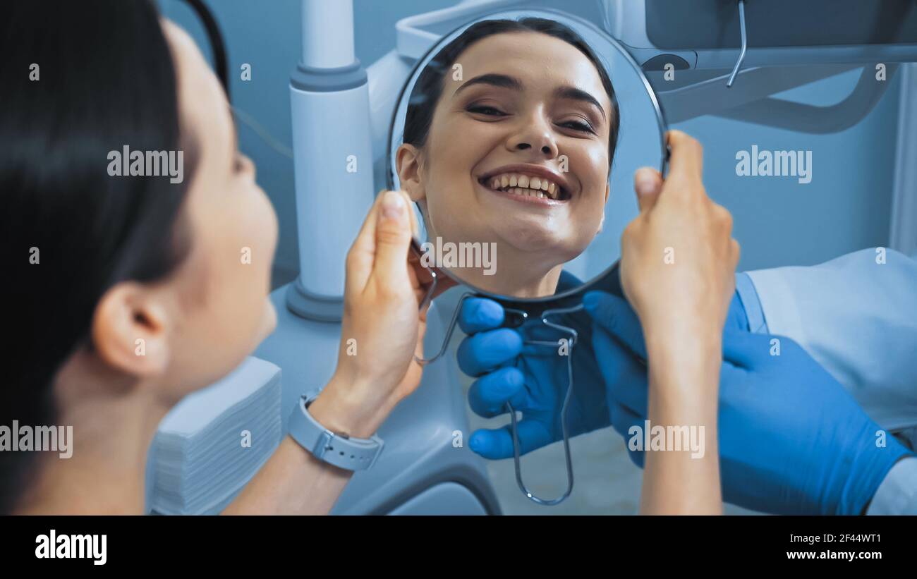 femme avec des dents parfaites regardant dans le miroir près de stomatologiste dans la clinique dentaire, flou de premier plan Banque D'Images