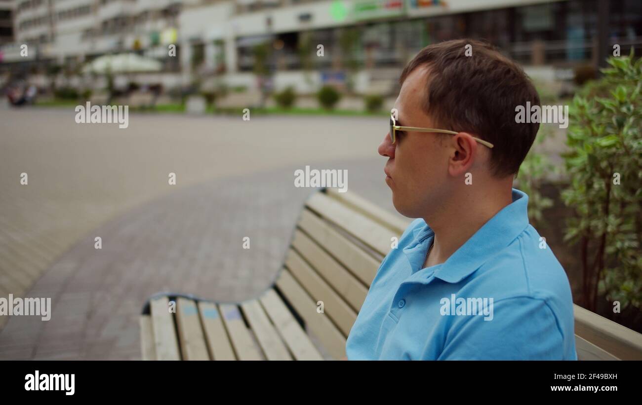 Homme dans des lunettes de soleil est de repos tout en étant assis sur un banc sur un fond flou de la ville, mouvement de caméra Banque D'Images