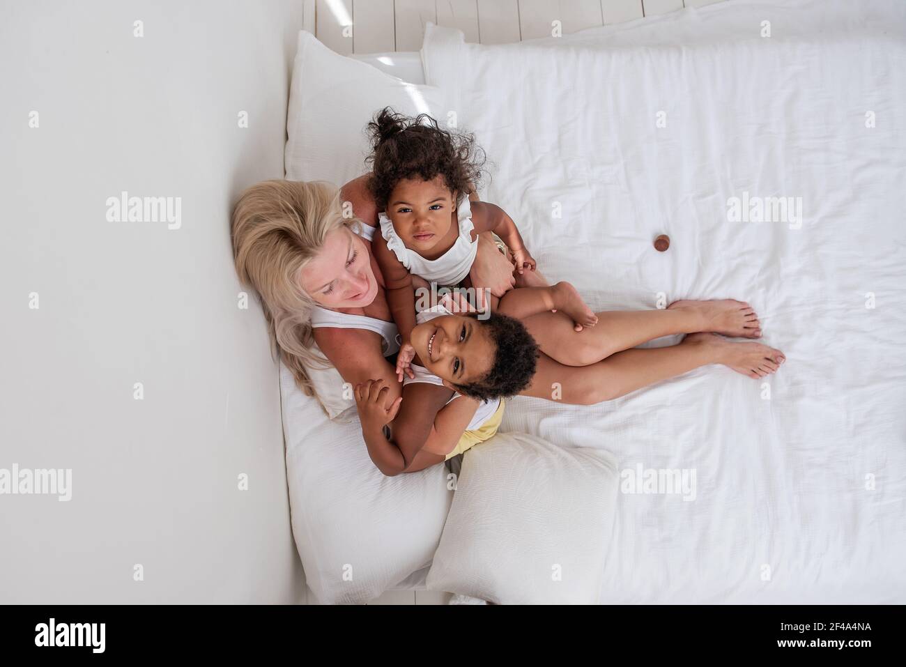 Un garçon et une fille afro-américains sont couchés sur un lit blanc, entre eux une jeune mère blanche blonde est assise. La femme embrasse la fille heureuse Banque D'Images