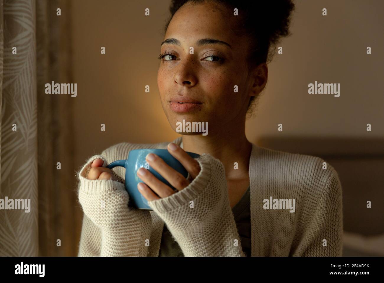 Femme de race mixte debout près d'une fenêtre et d'une tasse de boisson de café à la maison Banque D'Images