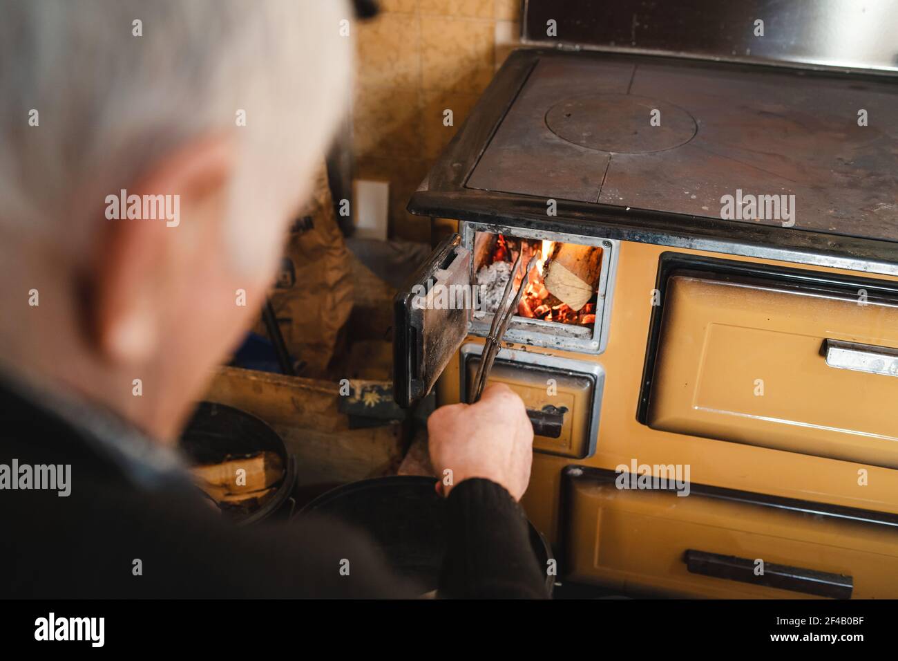 vue arrière en haut de l'homme âgé grand-père paysan caucasien assis près de la cuisinière de cuisine à sa maison à ferme mettant des bois sur la cheminée Banque D'Images