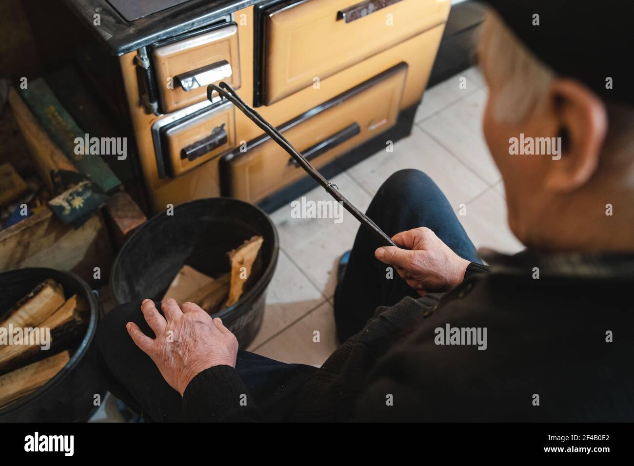 vue arrière en haut de l'homme âgé grand-père paysan caucasien assis près de la cuisinière de cuisine à sa maison à ferme mettant des bois sur la cheminée Banque D'Images
