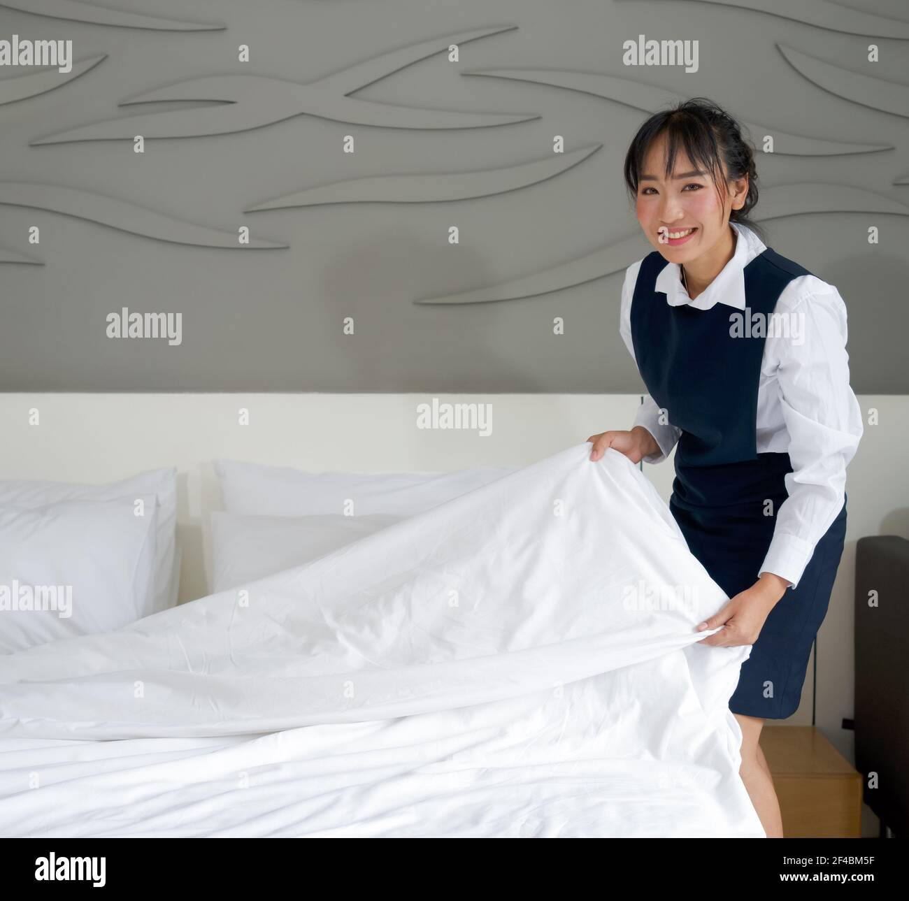 Jeune femme de chambre d'hôtel avec un sourire faisant le lit. Personnel en uniforme bleu préparant la chambre pour les clients de l'hôtel. Banque D'Images