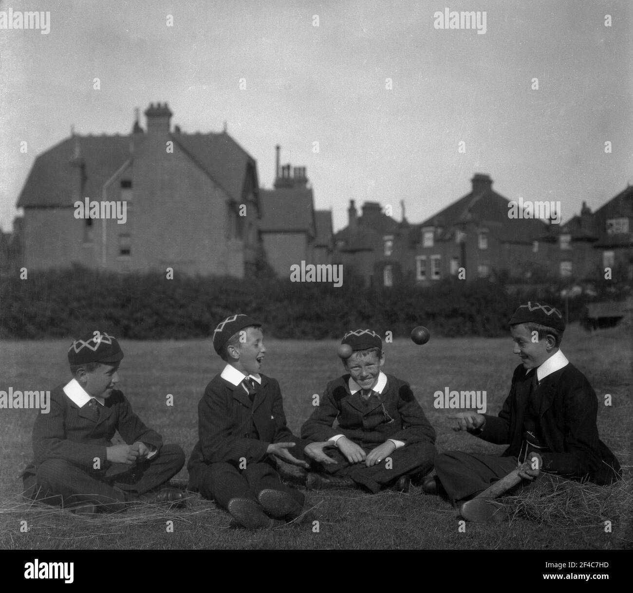 1930s, historique, assis ensemble dans un champ à l'extérieur, quatre garçons d'école publique anglaise dans leur uniforme et casquettes traditionnels, jouant 'catch' avec un ballon de cricket, Angleterre, Royaume-Uni. Banque D'Images
