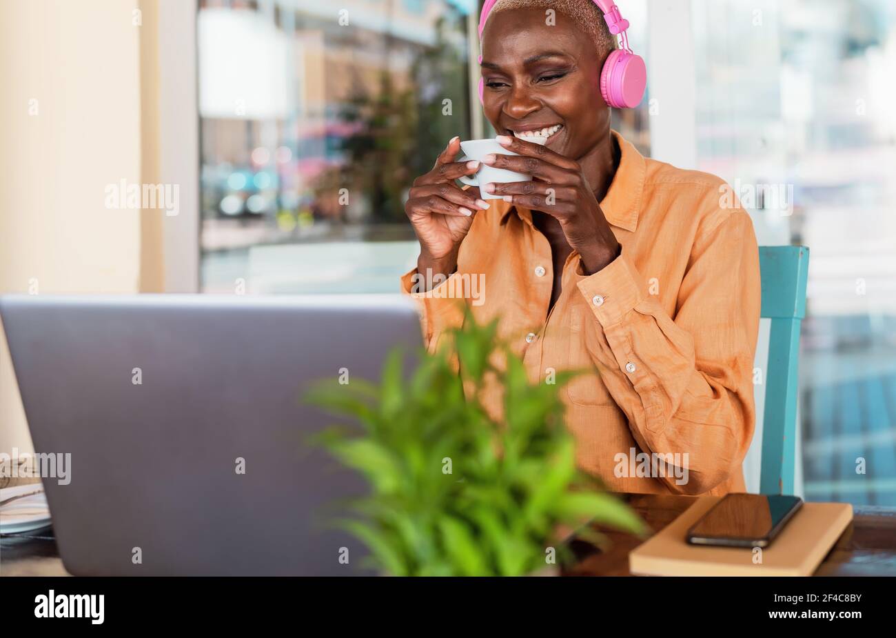 Femme africaine souriante qui boit du café tout en utilisant un ordinateur portable et en écoutant musique avec écouteurs dans le bar-restaurant Banque D'Images