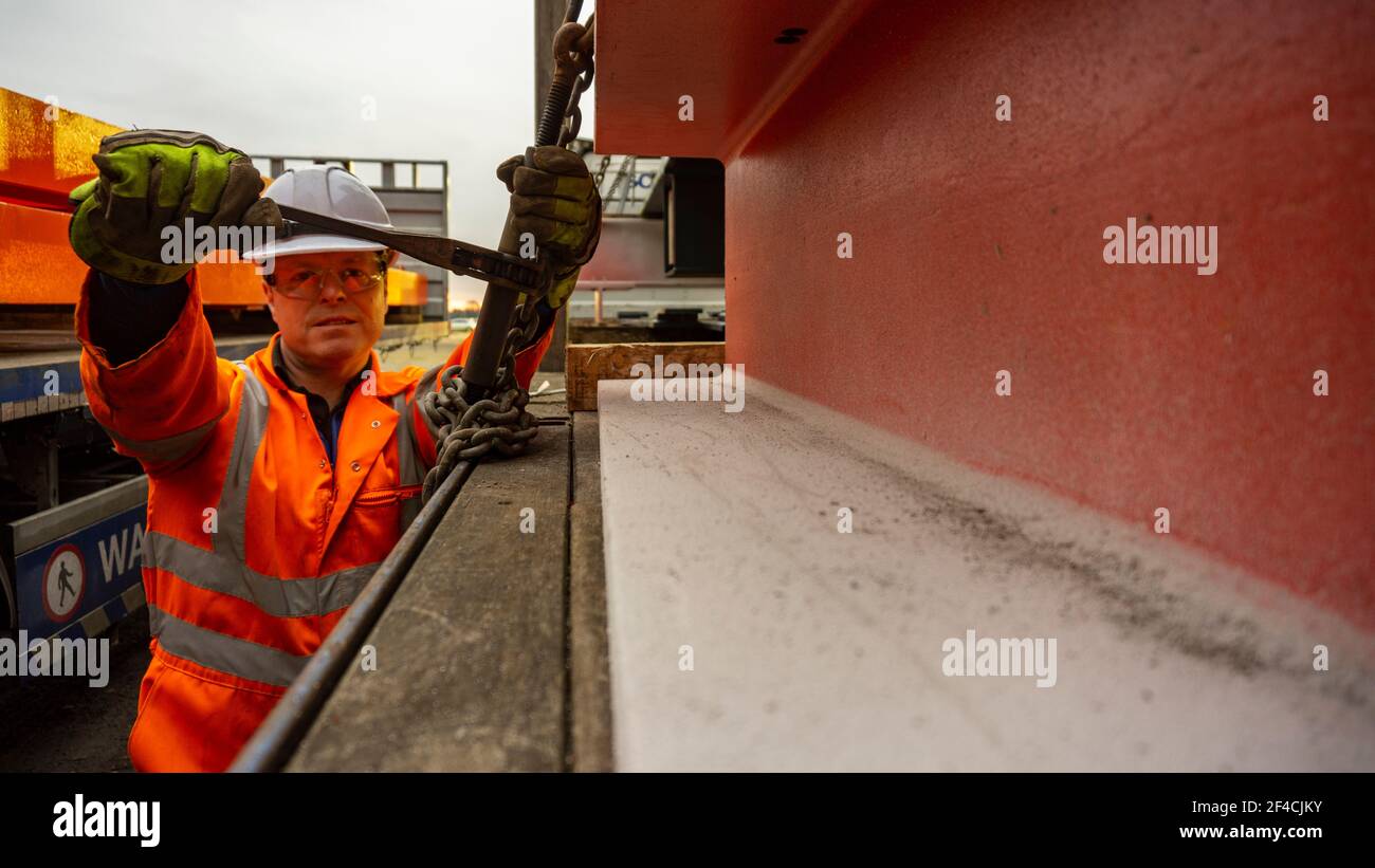 Conducteur de sexe masculin portant des combinaisons, des lunettes de protection, un casque et des gants de protection orange, qui portent un équipement de protection individuelle complet, qui fixe une charge d'acier sur une remorque à plateau. Banque D'Images