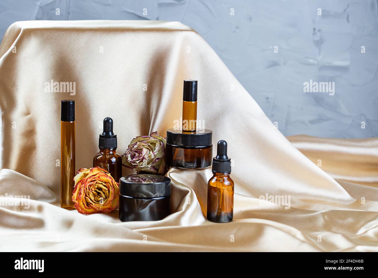Divers contenants pour le stockage de cosmétiques en verre foncé se trouvent sur les plis de tissu satiné de soie, entouré de fleurs de rose séchées. Parfum, sérum ou Banque D'Images