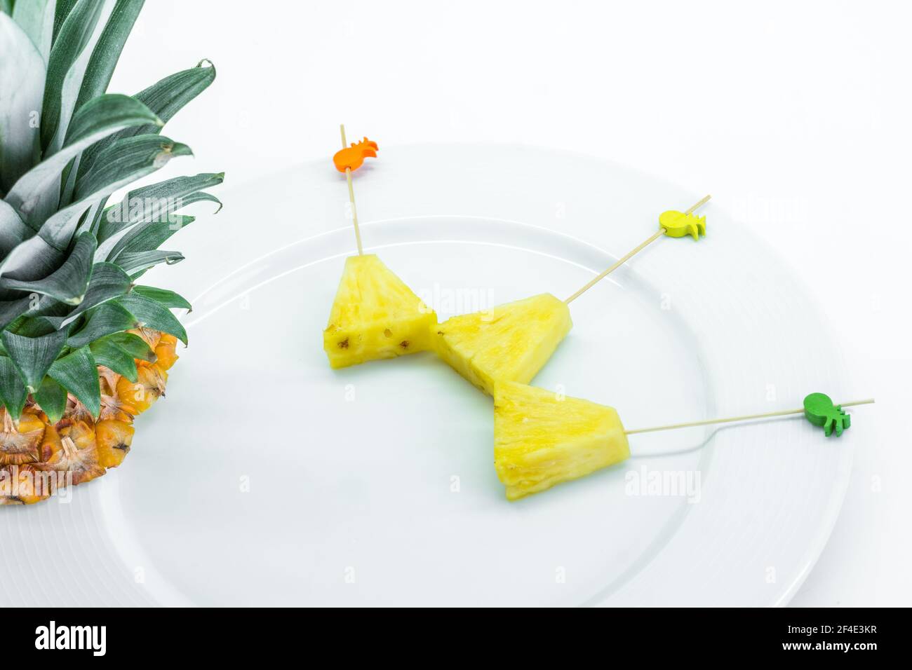 Brochettes d'ananas épluchées et coupées en triangles avec baguettes décorées sur plaque blanche avec feuilles d'ananas sur fond blanc Banque D'Images