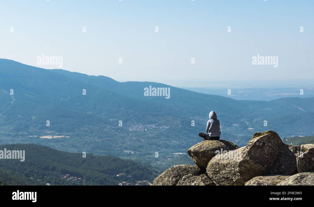 Fille dans un sweat à capuche gris assis sur un rocher de une falaise qui regarde le paysage de montagne s'étendant jusqu'au horizon Banque D'Images