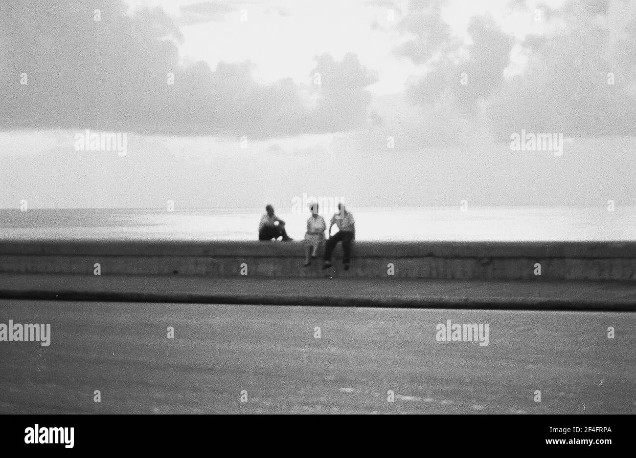 Gros plan de deux hommes et d'une femme assis sur un bas sentier côtier, avec l'océan en arrière-plan, sur le Malecon (Avenida de Maceo), la Havane, Cuba, 1964. () Banque D'Images